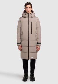 Lang, beige pufferjakke med lynlåslukning foran, justerbar hætte, lynlåslomme på brystet og sidelommer; har sorte detaljer ved ærmerne.