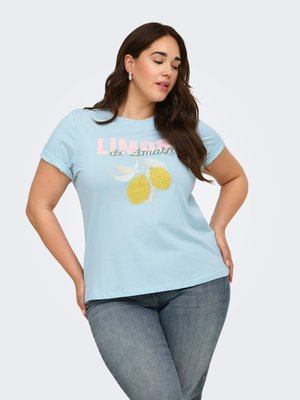 Femme posant les mains sur les hanches portant un t-shirt bleu clair avec des citrons et le texte « Limoni di Amalfi », assorti à un jean bleu.