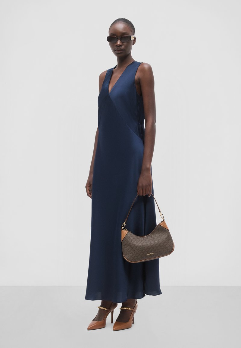 Femme portant une robe longue bleu marine sans manches, des talons pointus marron, des lunettes de soleil foncées, et tenant un sac à main marron avec des accents beige.
