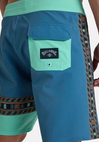 Pantalones de baño azul y turquesa con un bolsillo trasero con un parche de logo, intrincados patrones laterales y una textura suave.