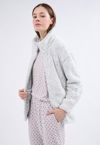 Chaqueta de piel sintética gris con cuello, que presenta una textura suave. Lleva puesta una camiseta gris clara con patrones de corazones rojos. La modelo está de pie con las manos en los bolsillos.