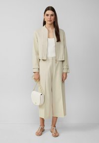 Beige cropped jasje over een witte top, gecombineerd met wijde beige broeken. Een crèmekleurige handtas met een kettinghandvat en tan sandalen maken de look compleet.