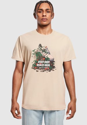 Beige t-shirt featuring a graphic of a sneaker surrounded by green vines and soil, with text reading "ROOT DEEP. REACH HIGH."