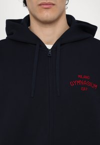 Marinblå zip-up hoodie med röd broderad text "MILANO GYMNASIUM EA7", med huva och justerbara dragsko. Mjuk textur.