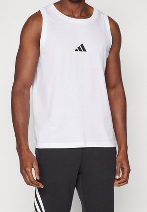 Homme portant un débardeur blanc sans manches avec un logo Adidas noir et un pantalon noir avec des bandes blanches sur les côtés, debout devant un fond uni.