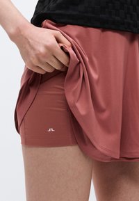 Mano levanta falda color rosa para revelar pantalones cortos integrados a juego con pequeño logo blanco en el muslo izquierdo.