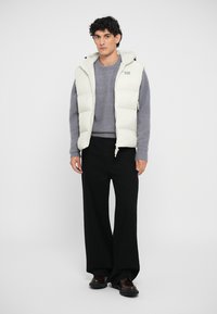 Puffer vest in off-white with a zippered front, adjustable hood, and a logo detail. Paired with a gray sweater and black wide-leg trousers.