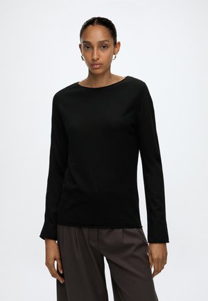 Vrouw met naar achteren getrokken haar, met gouden oorringen, een zwart shirt met lange mouwen en losse donkerbruine broek, staand tegen een effen achtergrond.