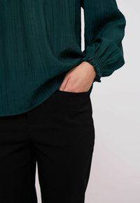 Teal tekstuuriga särk, millel on volditud varrukad, on sisse pandud mustadesse kitsastes pantidesse, käsi on pükste taskus.