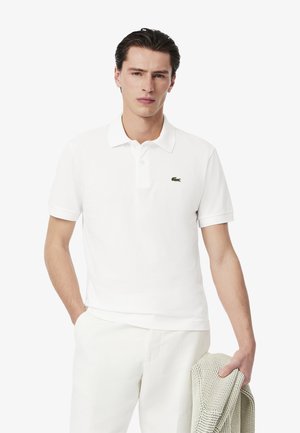 Camisa polo blanca de algodón con cuello tipo solapa, mangas cortas y un logo de cocodrilo bordado en verde en el lado izquierdo del pecho.