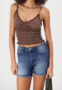 Femme portant un haut court brun plissé à fines bretelles et un short en denim bleu taille haute, tenant un tissu vert dans sa main droite.