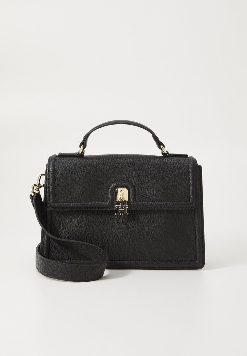 Sac à main en cuir noir avec poignée supérieure, fermoir doré présentant des lettres entrelacées, et bandoulière réglable reposant devant.