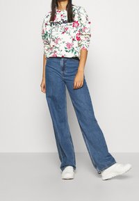 Sudadera color crema con estampado floral y logo negro, combinada con vaqueros de talle alto y pierna ancha en azul y zapatillas blancas.