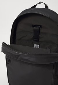 Svart nylon ryggsekk med glidelås, med flere interne lommer og en etikett. Glatt tekstur og minimalistisk design.