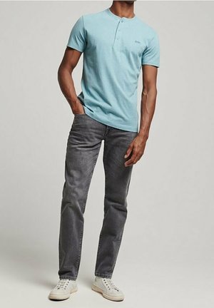 Mann trägt hellblaues Henley-Shirt mit kurzen Ärmeln, dunkelgraue Jeans und weiße Sneakers, steht mit einer Hand in der Tasche vor einem einfarbigen Hintergrund.