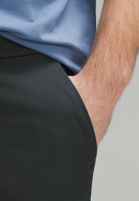 Czarne materiałowe spodnie o gładkiej i eleganckiej teksturze, z boczną kieszenią i częściowo widoczną jasnoniebieską koszulą w talii.