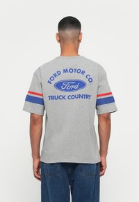 Siva pamučna majica s kratkim rukavima, s plavim i crvenim prugama na rukavima i velikim plavim Ford logotipom s tekstom na leđima.
