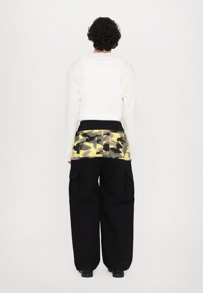 O bluză albă cu mâneci lungi, purtată împreună cu pantaloni cargo negri, având o bandă cu imprimeu camuflaj galben și verde la talie și buzunare vizibile pe pantaloni.