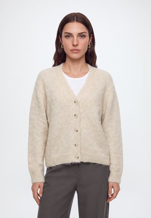 Femme aux cheveux bruns raides porte un cardigan crème boutonné sur une chemise blanche et un pantalon gris, debout devant un fond blanc uni.