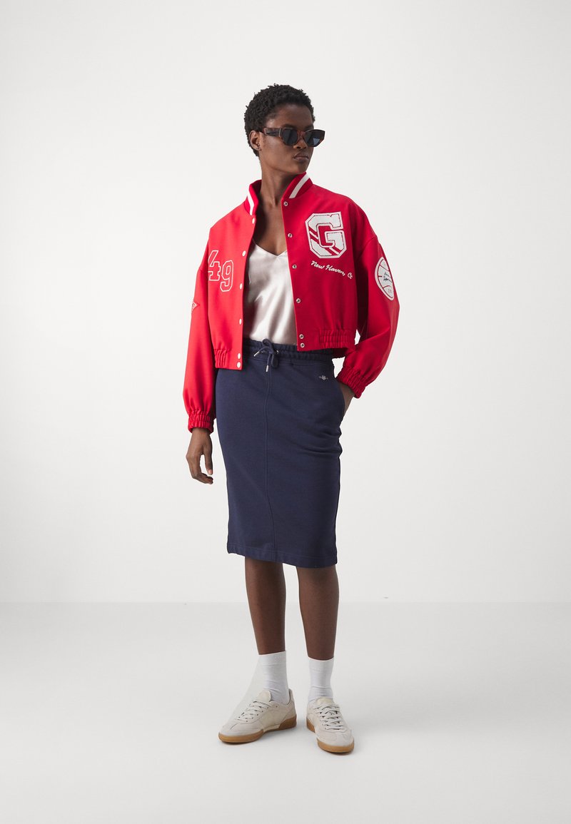 Czerwona krótka kurtka varsity z wyszywanym logo, długimi rękawami. Biała satynowa bluzka, granatowa spódnica ołówkowa i jasne sneakersy z beżowymi podeszwami.