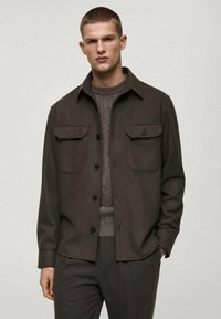 Dark brown shirt jacket made of soft fabric, featuring two chest pockets, a button front closure, and a classic collar. Worn over a textured sweater.