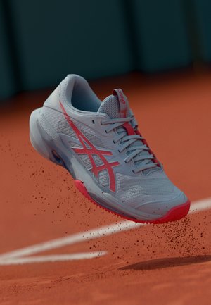 Lichtgrijze tennis schoen met roze accenten, met een mesh bovenwerk, gestructureerde zool en een onderscheidend logo, boven een klei tennisbaan zwevend.