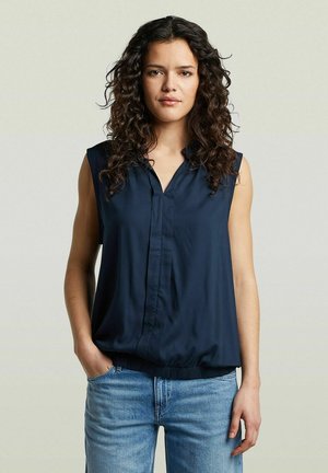 Femme aux longs cheveux bouclés portant un chemisier sans manches bleu marine et un jean bleu, debout avec une main dans la poche devant un fond uni.