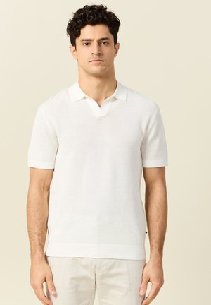 Mann trägt kurzärmliges, weißes, strukturiertes Polohemd und helle Hose, steht vor einfarbigem beigem Hintergrund.