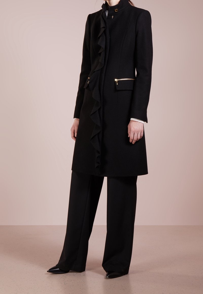 Cappotto di lana nero con dettagli arricciati sulla parte anteriore, finiture in oro e tasche laterali. Abbinato a pantaloni neri a gamba larga e scarpe a punta.