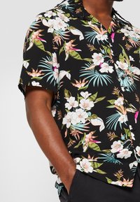 Camicia nera a maniche corte con bottoni, decorata con una stampa floreale tropicale nei colori rosa, bianco e verde, indossata da una persona con una mano nella tasca.