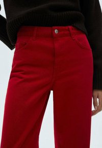 Rode denim broek met een slim fit, voorzakken en een metalen knoopsluiting. Gecombineerd met een donkerbruine gebreide trui.