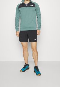Uomo che indossa un pullover teal con mezza zip, pantaloni corti neri e scarpe da corsa su trail blu, in piedi su un pavimento grigio chiaro contro un muro bianco.