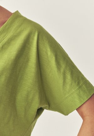 Lähikuva henkilön käsivarresta, jossa on päällä vaaleanvihreä, lyhythihainen puuvillapaita yksiväristä off-white-taustaa vasten.