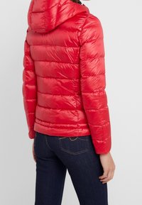 Rode gewatteerde jas met capuchon, voorzien van doorgestikte secties, glad glanzend oppervlak en aansluitend ontwerp. Gedragen over donkerblauwe spijkerbroek.