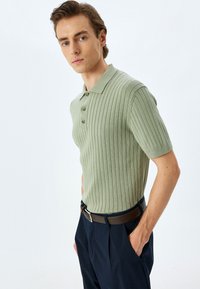 Ljust grön rippad polo-shirt med krage, korta ärmar, tre-knapps knäppning, bärs med mörka byxor, komplementerat med ett brunt bälte.