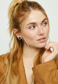 Pendientes de botón con piedras preciosas verdes, montados en plata, con forma redonda y superficie lisa, usados por una persona de cabello largo y rubio.