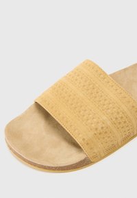 Gul semsket sklisandaler med en bred reim som har teksturerte utskjæringsmønstre, en glatt såle og en brun gummisole.
