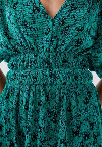 Robe verte à motifs avec décolleté en V, taille élastique et tissu texturé présentant des rayures verticales et des accents noirs.