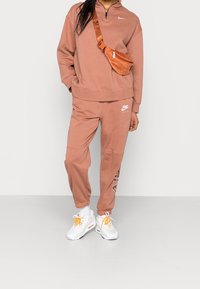 Rosa-brun Nike sweatshirt med halvdragkedja och logotyp, tillsammans med matchande joggers med logotyp och vita sneakers med orange accenter.