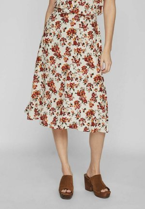 Frau, die einen weißen Midirock mit rot-orangefarbenem Blumenmuster und braunen Wildleder-Plateausandalen trägt, steht vor einem grauen Hintergrund.