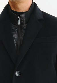 Czarny płaszcz z wełny z zamkiem błyskawicznym przy kołnierzu, z gładką satynową podszewką, z pojedynczym zapięciem na guzik i subtelnym detalem kieszeni.