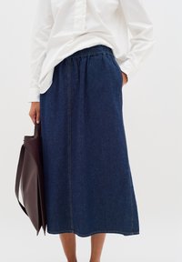 Jeanskjol med elastisk midjeband, mörkblå färg, knälång och synliga sömnadsdetaljer. Kombinerad med en vit långärmad skjorta och en brun väska.