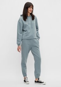 Hanorac albastru deschis și pantaloni jogger asortați, realizati din țesătură moale. Include o glugă cu șnur și manșete elastice. Asortați cu adidași negri din pânză.