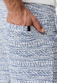 Pantalones cortos de tela ligera y texturizada con un patrón geométrico en blanco y azul, que cuentan con un bolsillo lateral con una etiqueta negra.