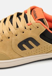 Tan színű velúr sneaker fekete hálós kiegészítőkkel, narancssárga részletekkel és gumitalppal. Jellemzője a kiemelkedő logó és a perforált dizájn elemek.