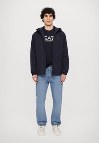 Marine blå vindjakke med hette, glidelås og elastiske mansjetter, kombinert med en marineblå T-skjorte og lyseblå jeans. Sneakers fullfører antrekket.
