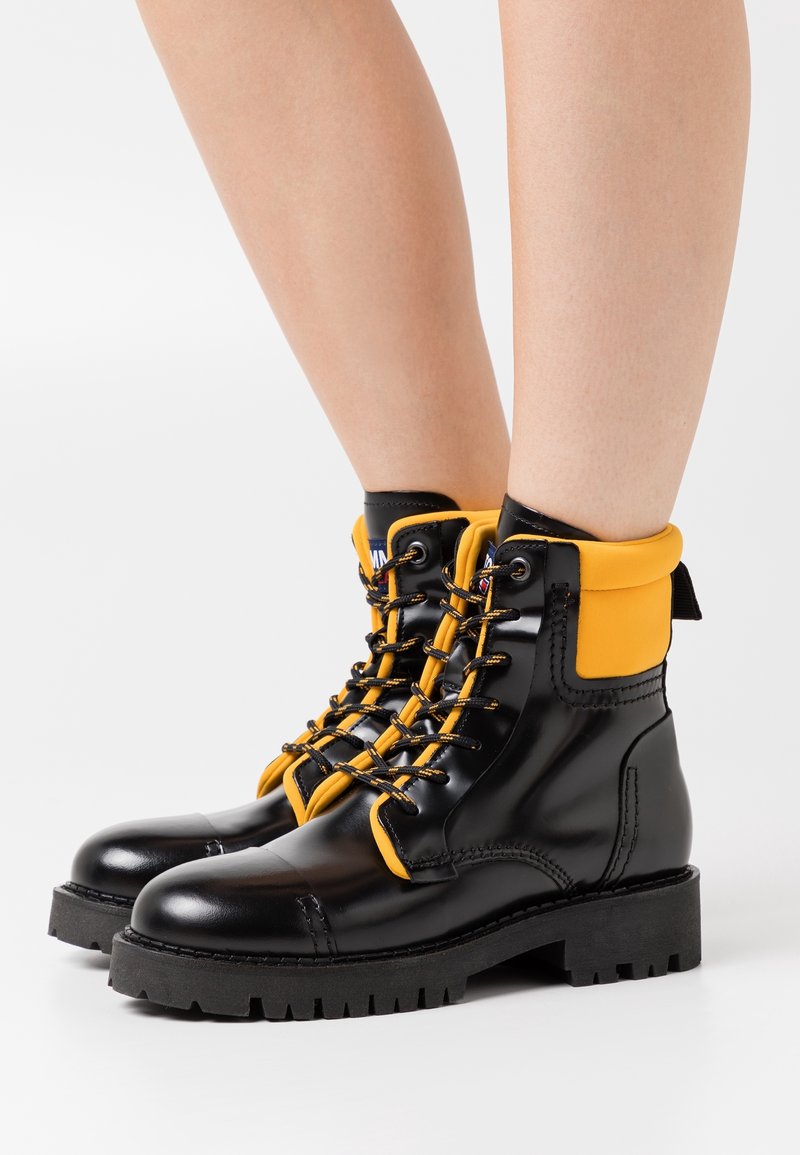 Bottes en cuir noir avec semelles épaisses, cols rembourrés jaunes et lacets noirs et jaunes portées sur des jambes nues.