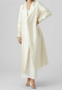 Dubbelt knäppt, lång kappa i glänsande vitt tyg med en slät yta, breda slag och sidofickor, bärs över en vit klänning.
