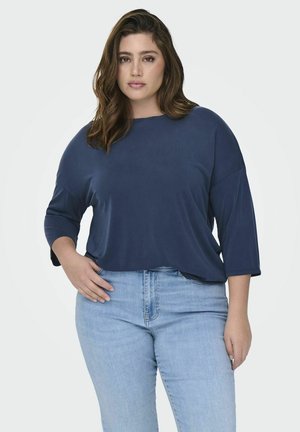 Marineblauwe, losse top met driekwartmouwen, gemaakt van zachte stof. Gepaard met lichtblauwe high-waisted jeans.