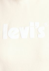 Tela color crema con letras blancas en relieve que forman la palabra "levi's®". La superficie es lisa, mostrando un diseño sencillo y una textura sutil.